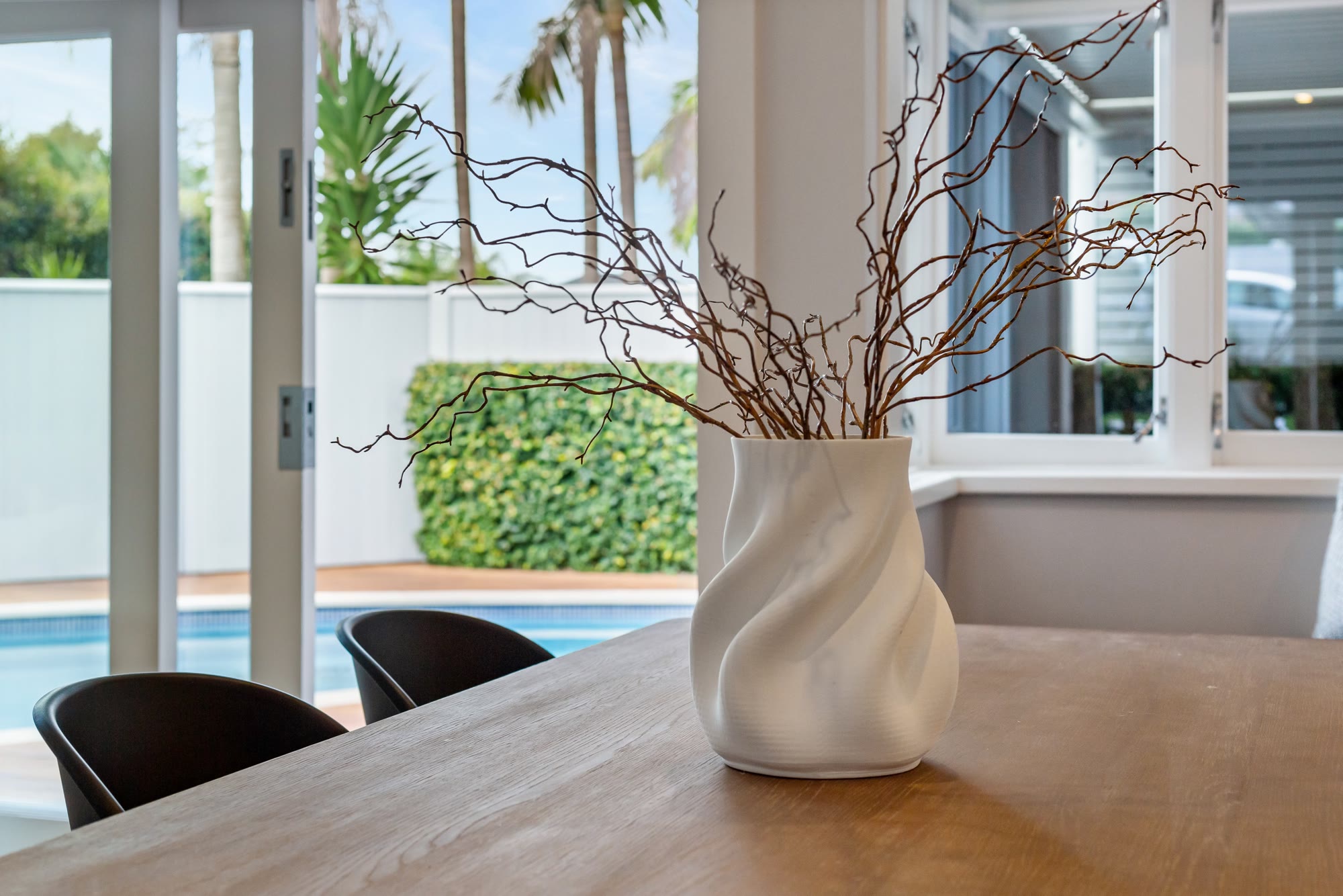 Timber dining table styled with a sculptural white vase beside a pool and garden outlook