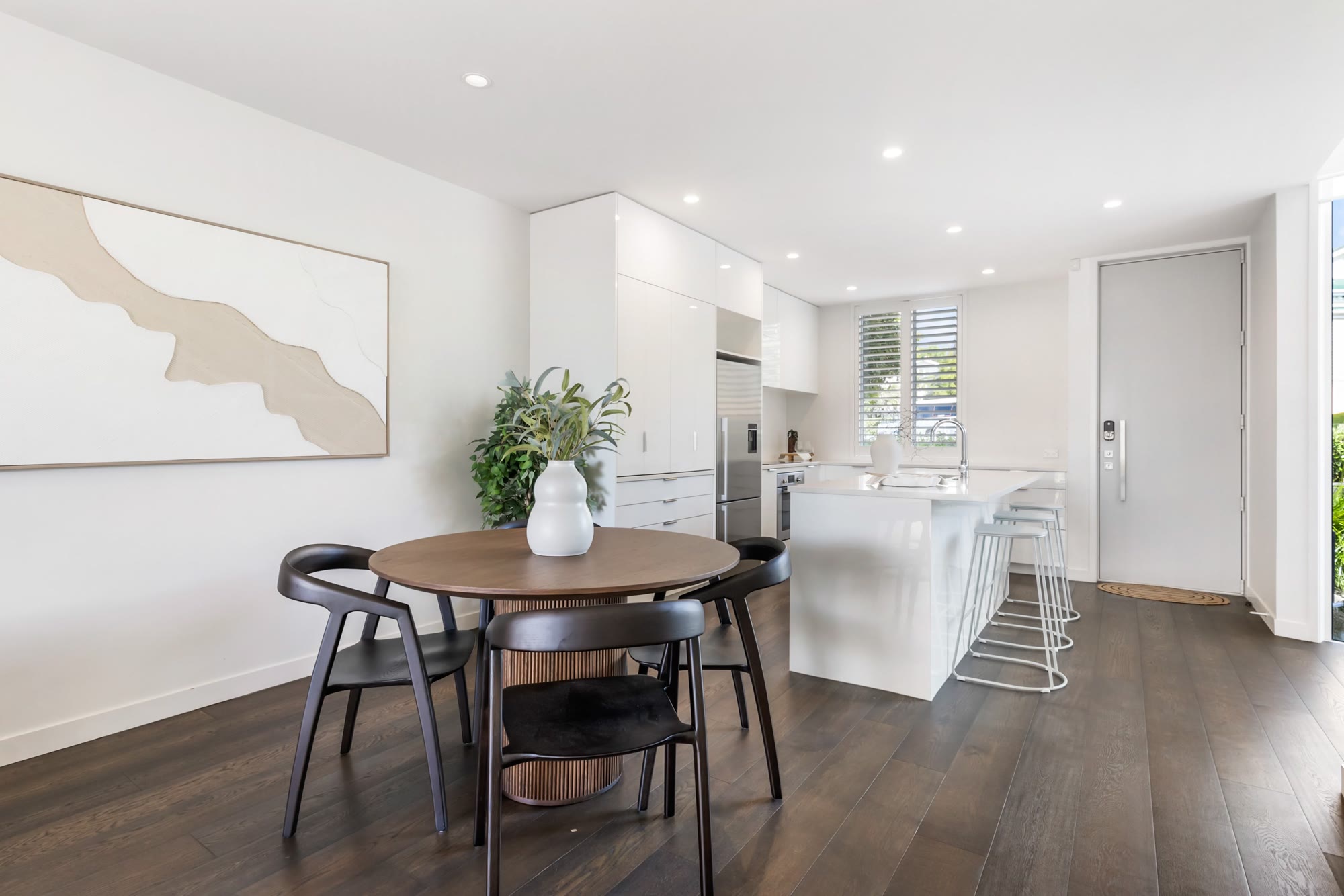 Round timber dining setting beside a bright white kitchen with abstract wall art