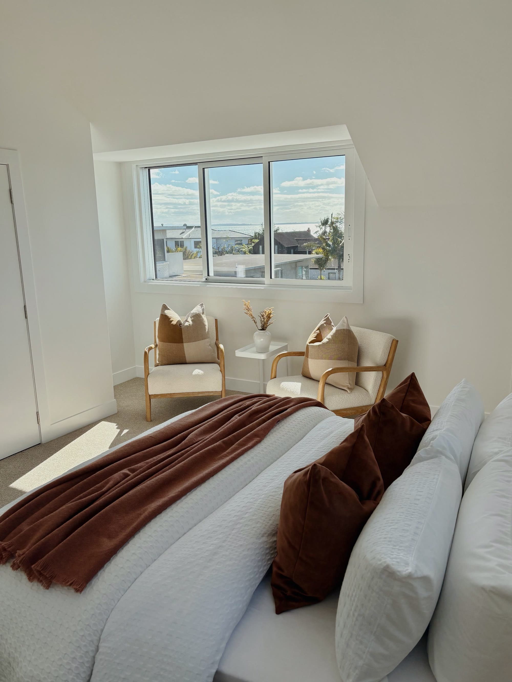 Sunlit bedroom with white bedding, rust cushions and a pair of timber occasional chairs