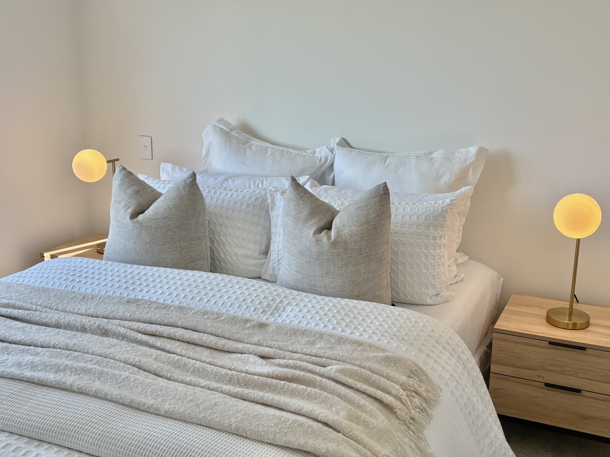 Bedroom styled with layered white bedding, soft grey cushions and matching warm bedside lamps