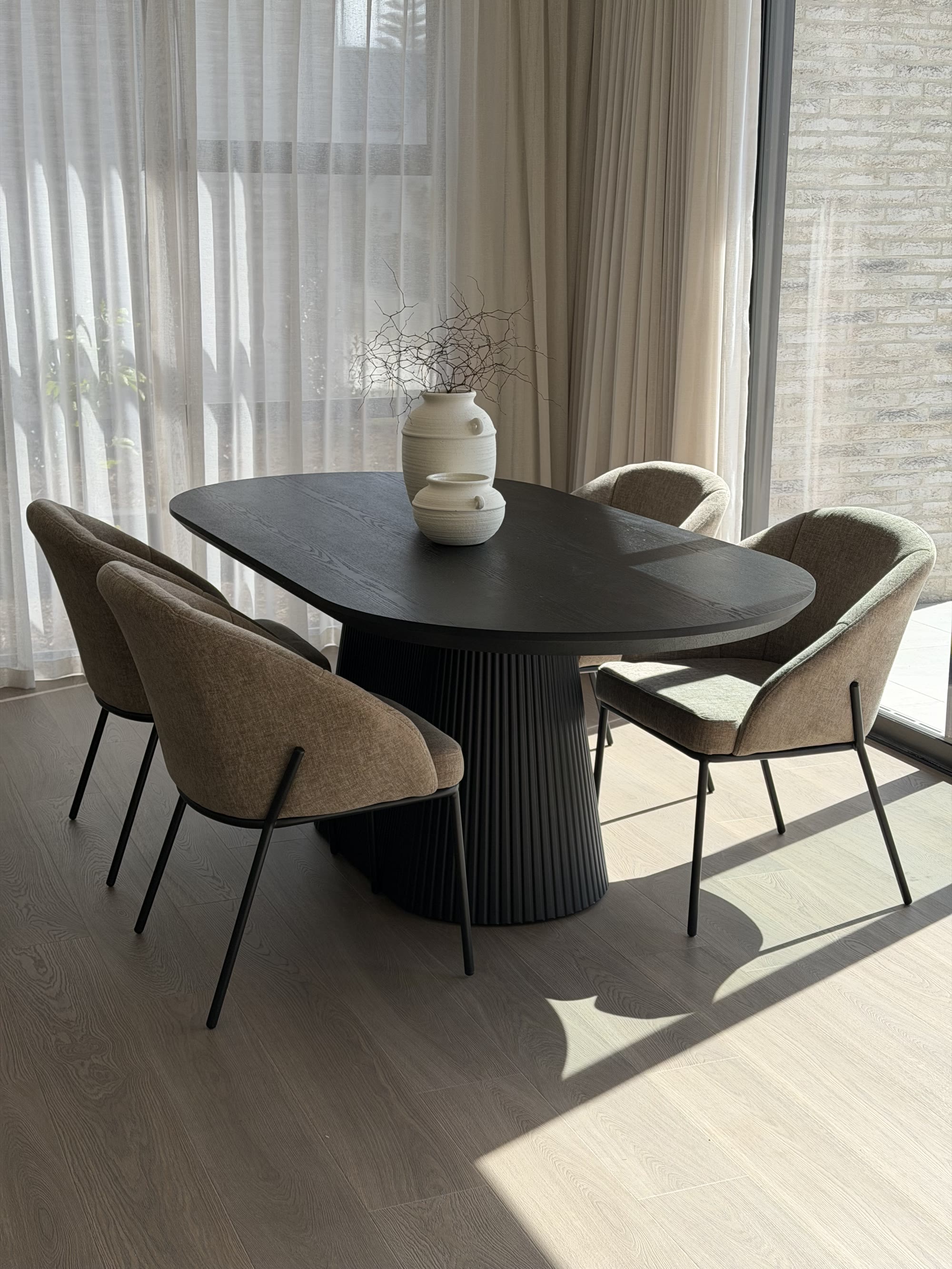 Black oval dining table with upholstered chairs and ceramic vases beside sheer curtains