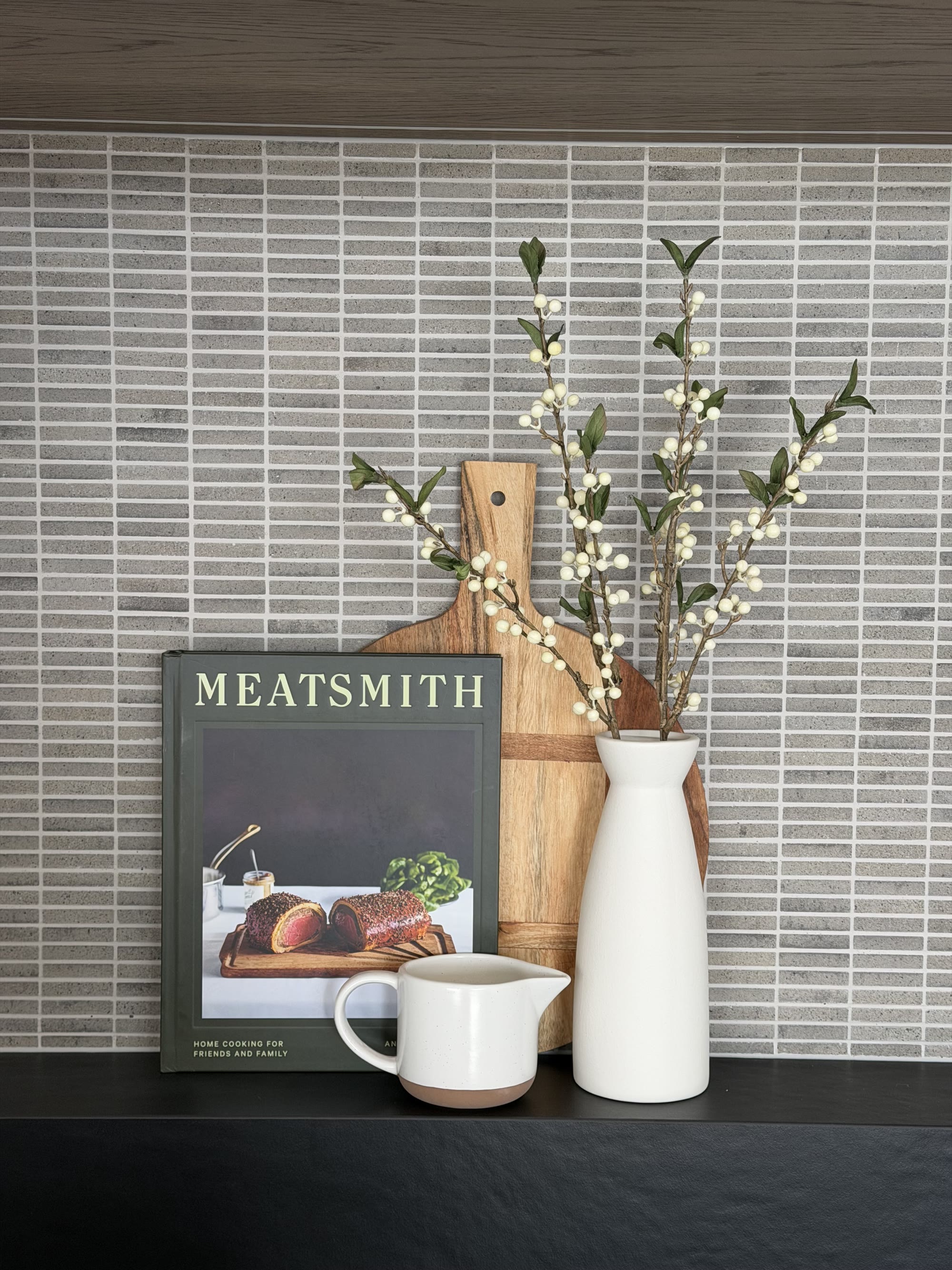 Kitchen bench styled with white ceramics, a timber board, cookbook and leafy stems