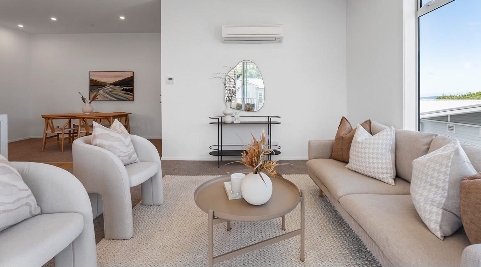 Open-plan apartment living and dining area with neutral seating, layered cushions and a coastal outlook